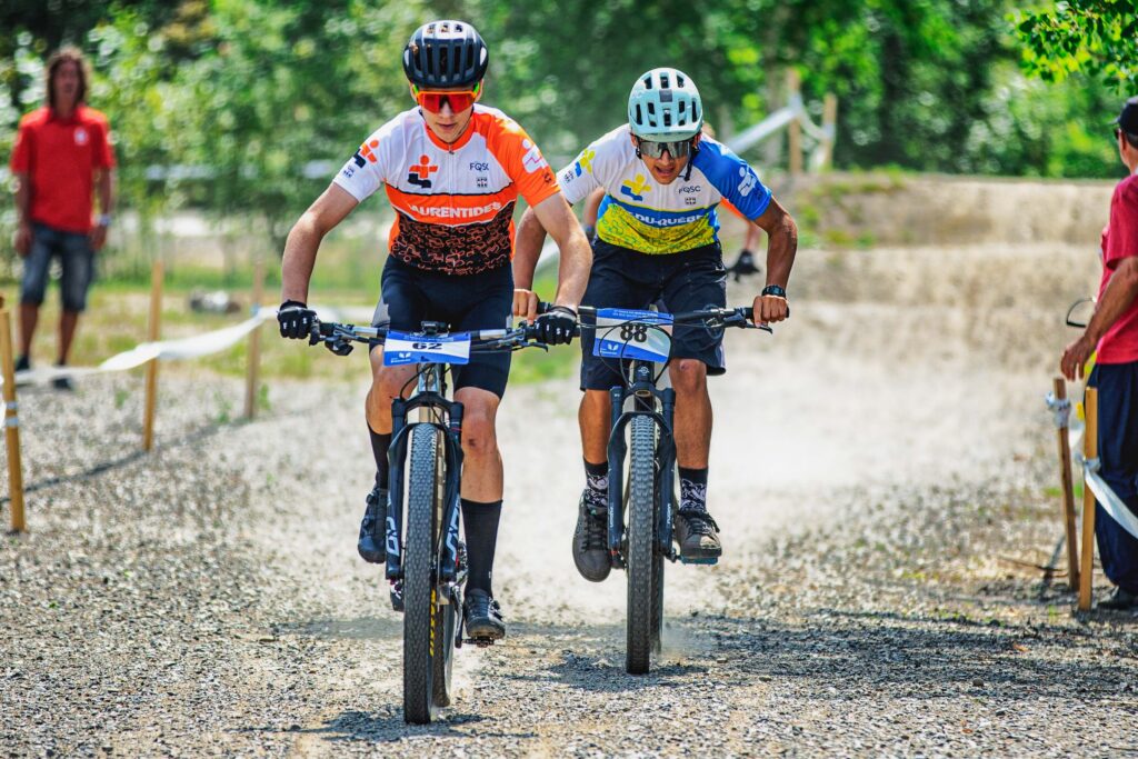 Deux cyclistes de vélo de montagne côte à côte en pleine course sur un sentier de gravier, portant des maillots régionaux.