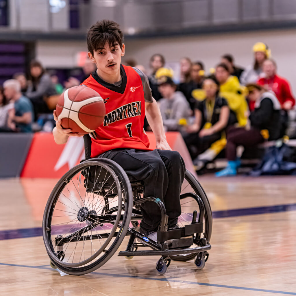 Un joueur de basketball en fauteuil roulant portant l'uniforme rouge de l’équipe de Montréal dribble un ballon sur le terrain, devant des spectateurs en arrière-plan.
